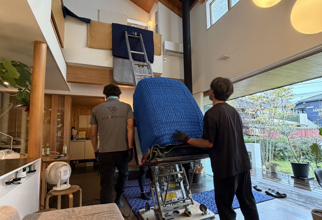 吹き抜けのある室内で、青い養生をした大型家具を二人がかりで慎重に運ぶ作業員